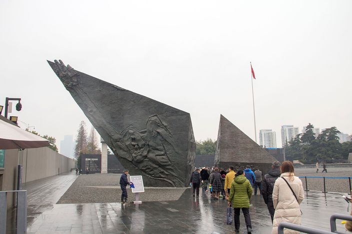 A large angular black stone sculpture with carved relief figures outside the Nanjing Massacre Memorial Hall, a red flag and visitors nearby under an overcast sky.