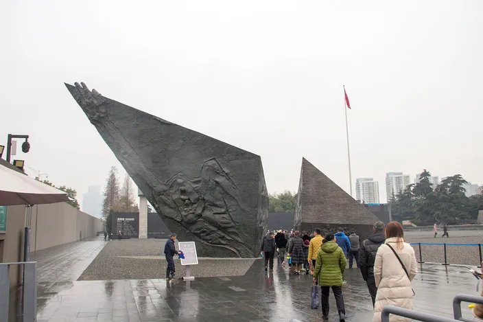 A large angular black stone sculpture with carved relief figures outside the Nanjing Massacre Memorial Hall, a red flag and visitors nearby under an overcast sky.