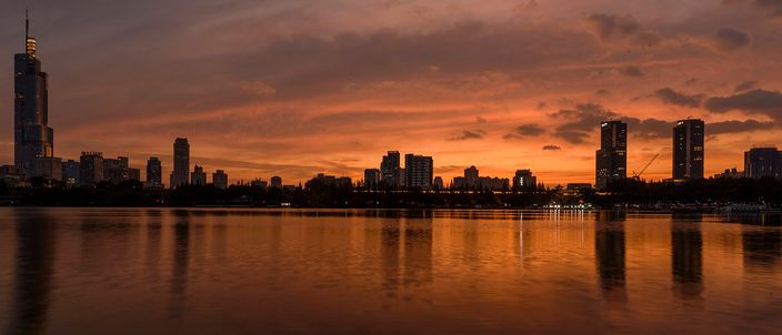 Nanjing's skyline reflected in Xuanwu Lake at dusk, the tapering Zifeng Tower silhouetted against an orange sky.