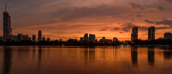 Nanjing's skyline reflected in Xuanwu Lake at dusk, the tapering Zifeng Tower silhouetted against an orange sky.