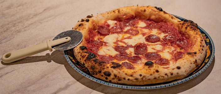 A Neapolitan pizza with salami on a decorated plate, its puffed rim blistered and leopard-spotted from the wood oven, a pizza cutter resting beside it.