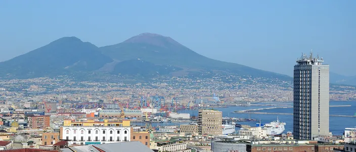 Daytime view over Naples rooftops to the twin-peaked cone of Vesuvius, with the port, a modern tower and the bay to the right under clear blue sky.