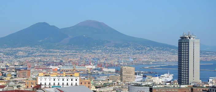 Daytime view over Naples rooftops to the twin-peaked cone of Vesuvius, with the port, a modern tower and the bay to the right under clear blue sky.