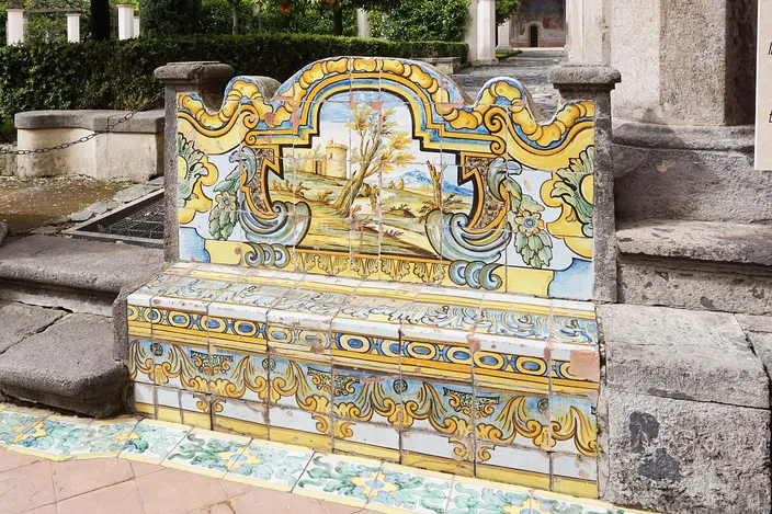 A long bench in a cloister walk, its back and seat entirely faced in painted majolica tiles — yellow scrollwork framing a panel of trees and a distant landscape — set against rough stone paving and a pillar, with the cloister garden behind.