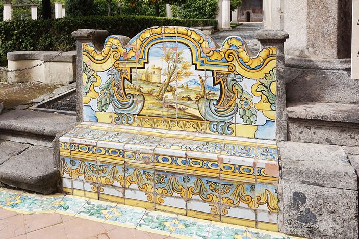 A long bench in a cloister walk, its back and seat entirely faced in painted majolica tiles — yellow scrollwork framing a panel of trees and a distant landscape — set against rough stone paving and a pillar, with the cloister garden behind.