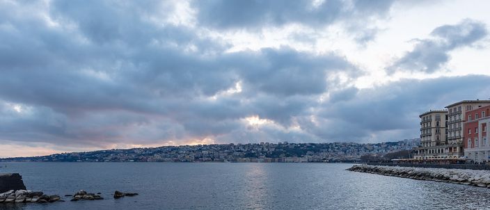 A wide dusk view along a curving seafront: dark calm water in the foreground, a low rock breakwater on the left, a city climbing a headland across the bay, and a large ornate palazzo on the promenade at the right under a heavy cloudy sky.