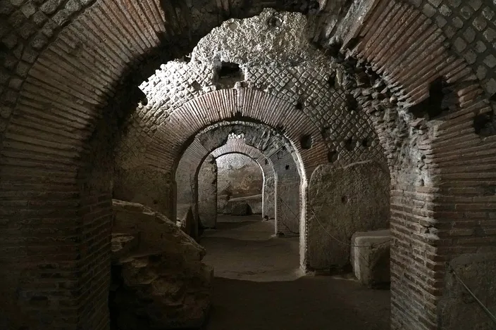 A dim underground corridor of Roman brick and rough stone: barrel vaults springing from square piers, an arched opening leading through to a further vaulted room, rubble and a cut stone block on the packed earth floor.