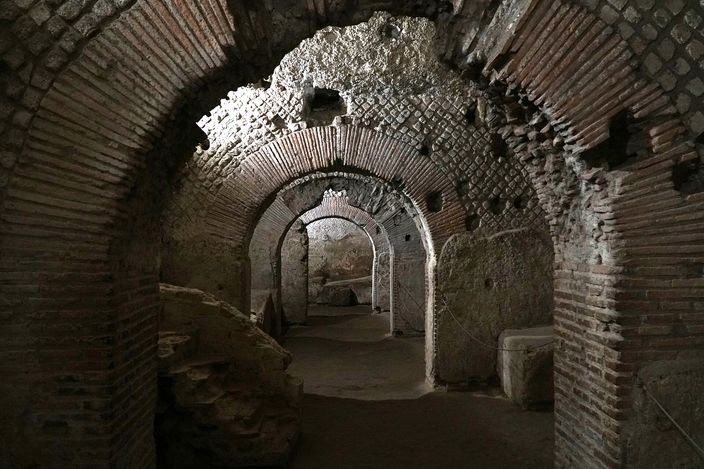 A dim underground corridor of Roman brick and rough stone: barrel vaults springing from square piers, an arched opening leading through to a further vaulted room, rubble and a cut stone block on the packed earth floor.