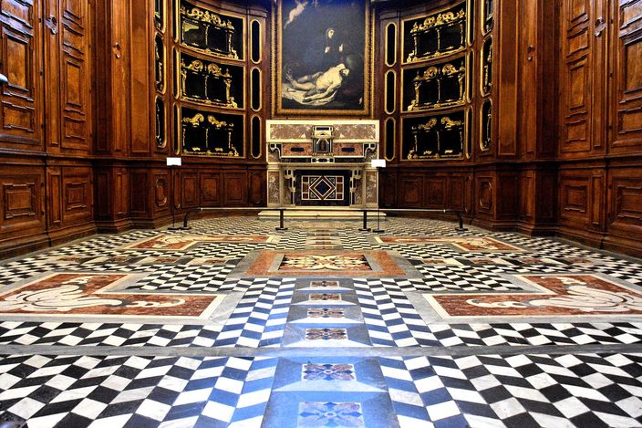 A wood-panelled apse-shaped room in a monastery: a dark oil painting of the dead Christ hangs above a marble altar set between tiers of inlaid panels, and the entire floor is a geometric pavement of white, black, blue and red inlaid marble running towards the viewer.