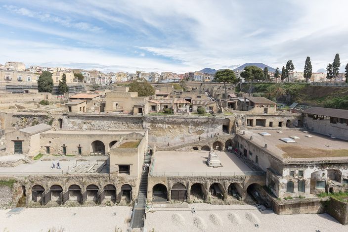 An excavated Roman town sunk in a deep rectangular pit: a row of arched brick vaults along the near edge, roofed and half-roofed houses on a grid of streets beyond, the modern town standing along the lip above, and the twin-peaked cone of Vesuvius on the horizon.