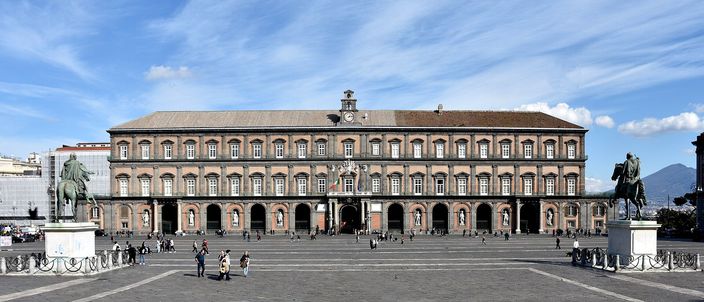 The long ochre facade of the Royal Palace fills the far side of Piazza del Plebiscito, its ground floor an arcade of deep arches; two bronze equestrian statues stand on tall plinths at either edge of the wide empty paved square, with a few people crossing it under a blue sky of white cloud.