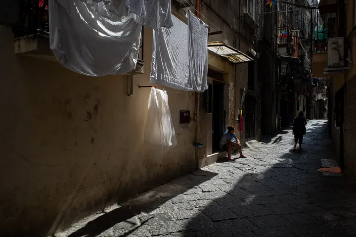 A narrow, sunlit stone-paved alley in the old town; white sheets hang to dry across the lane and a person sits on a step in the shadowed doorway of a worn building