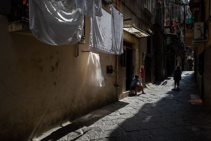 A narrow, sunlit stone-paved alley in the old town; white sheets hang to dry across the lane and a person sits on a step in the shadowed doorway of a worn building