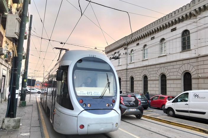 A modern white AnsaldoBreda Sirio tram, route 412, on street tracks beside a long classical stone building in Naples, with parked cars and overhead wires under a pink dawn sky