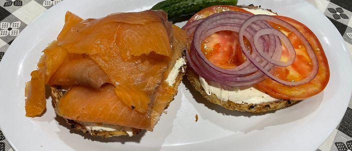 An open bagel on a diner plate: one half piled with cured salmon, the other with cream cheese, sliced tomato, red onion and a pickle.