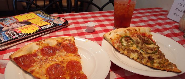 Two triangular slices of New York-style pizza on white plates on a red-and-white checked tablecloth, one topped with pepperoni, the thin crust visibly foldable.