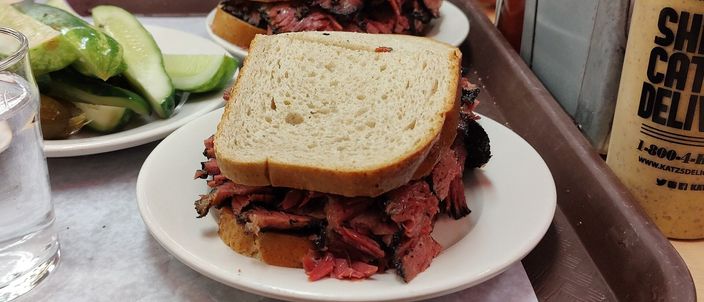 A pastrami on rye sandwich cut in half and stacked high with thick hand-carved slices of dark, peppery cured beef between two thin slices of rye bread, on a deli tray with a pickle.