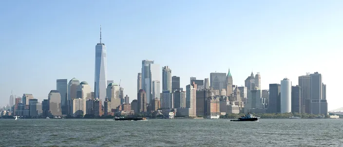 The Lower Manhattan skyline seen across Upper New York Bay on a clear day, One World Trade Center rising above the cluster of towers, with flat open water in the foreground.