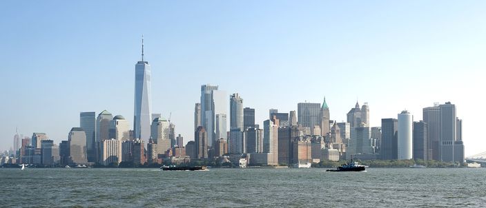 The Lower Manhattan skyline seen across Upper New York Bay on a clear day, One World Trade Center rising above the cluster of towers, with flat open water in the foreground.