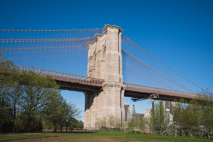The Brooklyn-side tower of the Brooklyn Bridge seen from the park beneath it: a pale stone pier with two pointed arches, suspension cables sweeping down on either side, trees and grass in the foreground and the roadway crossing behind.