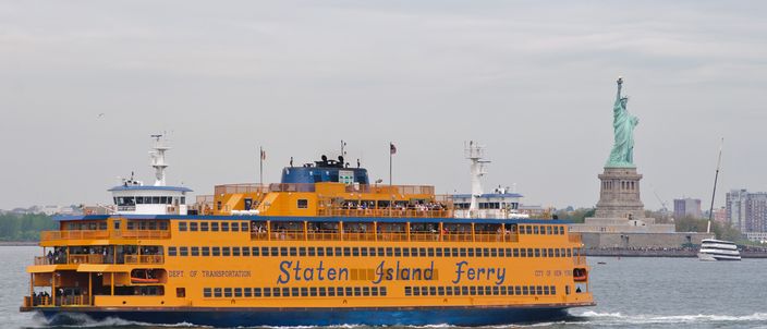 The orange Staten Island Ferry crossing the harbour in bright light, its name painted along the hull, with the Statue of Liberty standing on the far side of the water behind it.