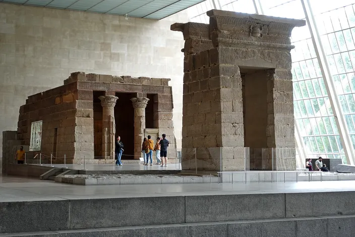 The sandstone Temple of Dendur reassembled indoors at the Metropolitan Museum, its gateway and shrine standing on a low plinth beside a shallow reflecting channel, visitors small against the stone and daylight falling through the sloping glass wall on the right.