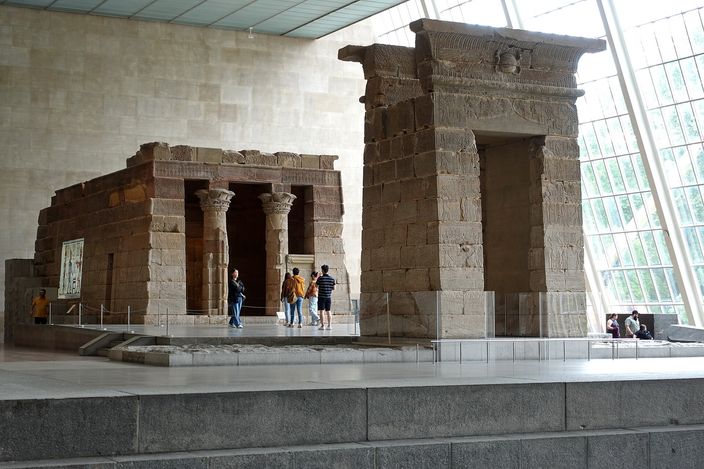 The sandstone Temple of Dendur reassembled indoors at the Metropolitan Museum, its gateway and shrine standing on a low plinth beside a shallow reflecting channel, visitors small against the stone and daylight falling through the sloping glass wall on the right.