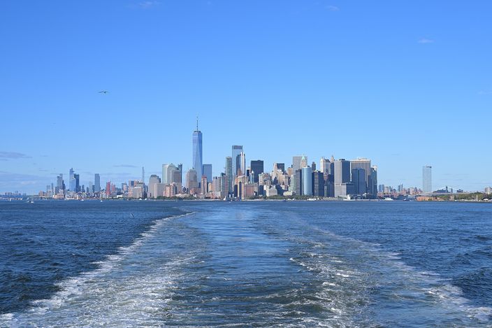 The Lower Manhattan skyline seen astern from a departing Staten Island Ferry, the boat's broad white wake filling the foreground water and the towers of the Financial District standing in a line on the horizon under a clear sky.