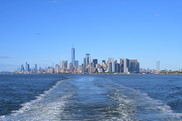 The Lower Manhattan skyline seen astern from a departing Staten Island Ferry, the boat's broad white wake filling the foreground water and the towers of the Financial District standing in a line on the horizon under a clear sky.