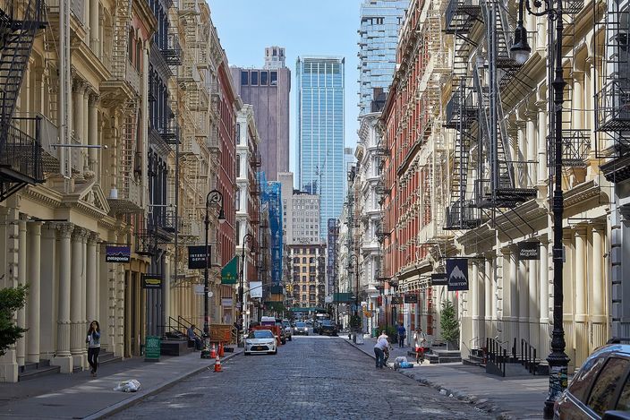 A cobbled block of Greene Street in SoHo lined on both sides with painted cast-iron warehouse fronts, columns and window surrounds repeating up the facades with fire escapes across them, the street angling away towards a modern tower at its far end.
