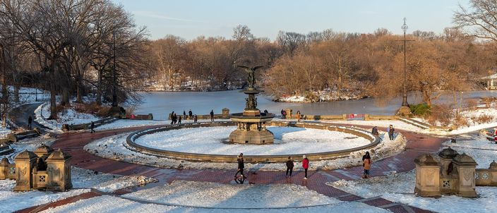 Snow lying on the red-brick paving and the rim of Bethesda Fountain in Central Park, the winged Angel of the Waters standing above the basin, walkers crossing the terrace and the part-frozen lake and bare woods behind.