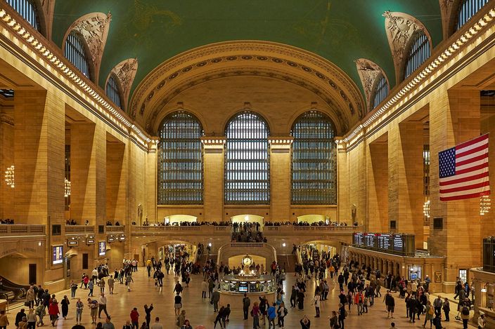 The main concourse of Grand Central Terminal, its vaulted green ceiling above tall arched windows, with commuters crossing the marble floor and an American flag hanging over them.