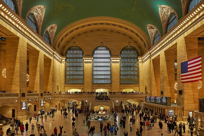 The main concourse of Grand Central Terminal, its vaulted green ceiling above tall arched windows, with commuters crossing the marble floor and an American flag hanging over them.