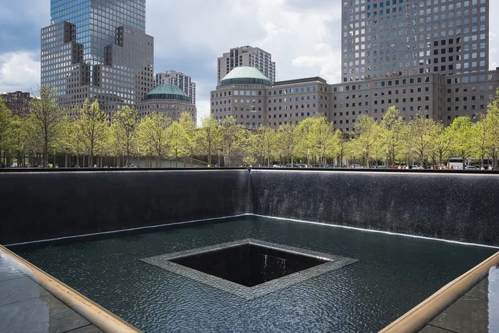 The north reflecting pool of the National September 11 Memorial: a square void with water falling down its black granite walls, edged by the bronze parapet carrying the names, with the surrounding towers and young trees behind.