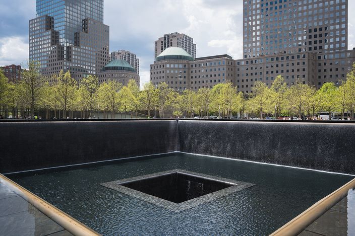 The north reflecting pool of the National September 11 Memorial: a square void with water falling down its black granite walls, edged by the bronze parapet carrying the names, with the surrounding towers and young trees behind.