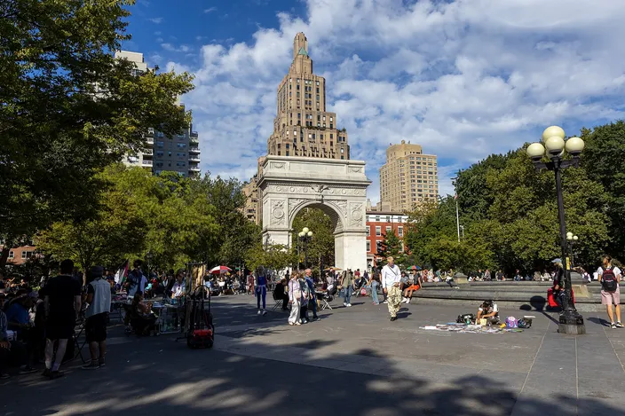 Washington Square Park on a summer afternoon: the marble arch at the north end, people sitting and walking on the paving around the fountain, trees on both sides and a tower behind.