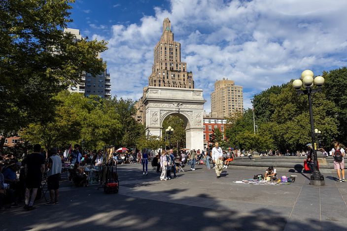 Washington Square Park on a summer afternoon: the marble arch at the north end, people sitting and walking on the paving around the fountain, trees on both sides and a tower behind.