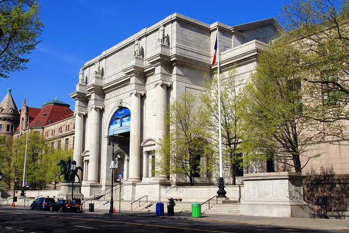 The columned entrance of the American Museum of Natural History behind bare spring trees, its pale stone facade and equestrian statue seen from the street corner under a clear sky.