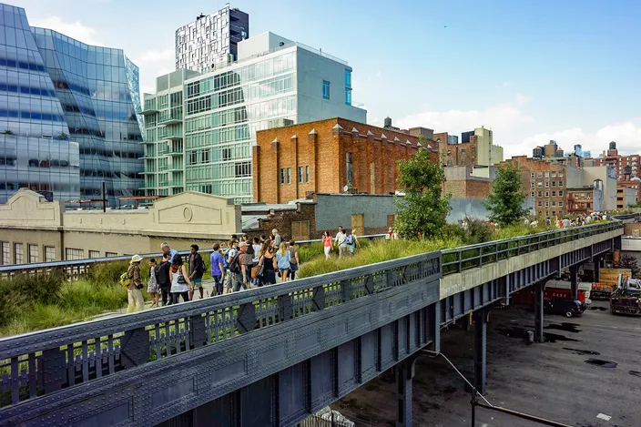 The High Line seen from street level: an elevated steel railway viaduct planted as a narrow park, people walking along the top of it between glass and brick buildings.