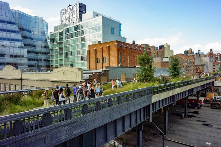 The High Line seen from street level: an elevated steel railway viaduct planted as a narrow park, people walking along the top of it between glass and brick buildings.
