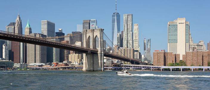 The Brooklyn Bridge crossing the East River in clear summer light, with the towers of Lower Manhattan behind it and a small boat passing underneath.