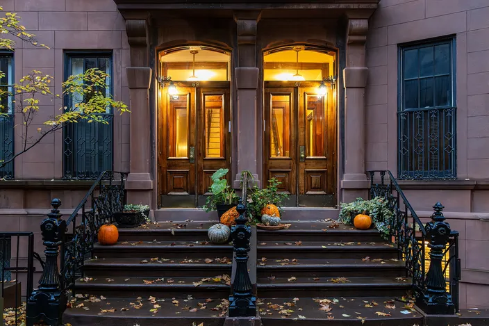 A Greenwich Village brownstone in autumn: a broad stone stoop with iron railings leads to a pair of tall lit doorways, pumpkins on the steps and fallen leaves at the curb.