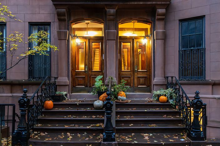 A Greenwich Village brownstone in autumn: a broad stone stoop with iron railings leads to a pair of tall lit doorways, pumpkins on the steps and fallen leaves at the kerb.