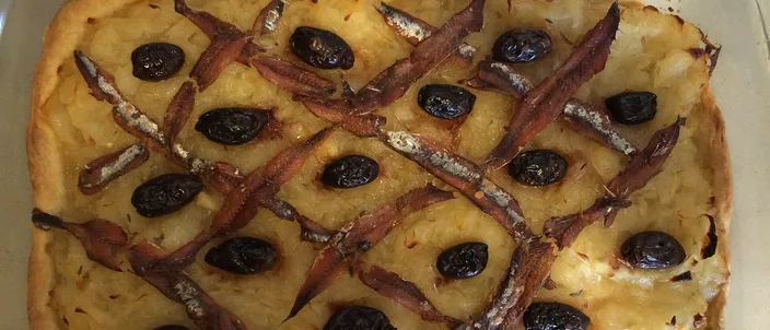 A rectangular pissaladière in a glass baking dish: pale, slow-cooked onions covered by a criss-cross lattice of anchovy fillets, with a black olive set in each diamond.