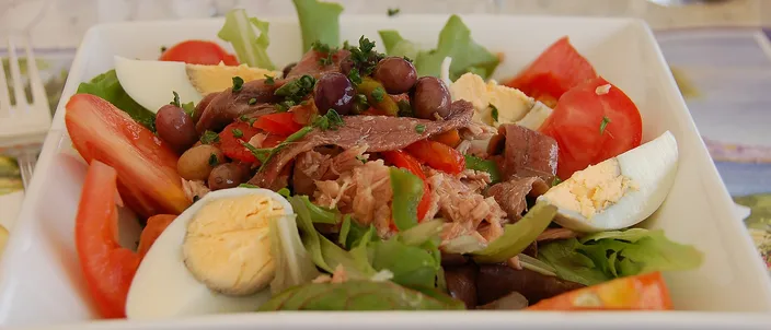 A salade niçoise in a white square bowl: wedges of red tomato, halved hard-boiled eggs, tuna, anchovy fillets, strips of green and red pepper and small black olives over a few leaves, with no potatoes or green beans.