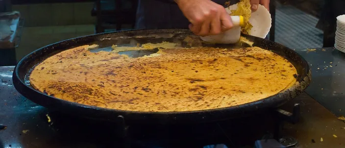 A cook at a counter in Nice scraping freshly baked socca off a huge round black tray with a spatula, the thin chickpea pancake golden and freckled with dark scorch marks.