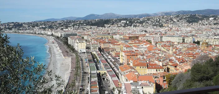 View from the castle hill over Nice: the Promenade des Anglais and pebble beach curving along the blue bay to the left, with the terracotta rooftops of Vieux-Nice spreading below and hills on the horizon.