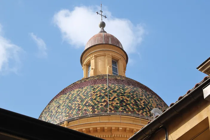 The dome of Nice's Cathédrale Sainte-Réparate, covered in green, yellow and red glazed tiles and topped by a small lantern and cross, against a blue sky.