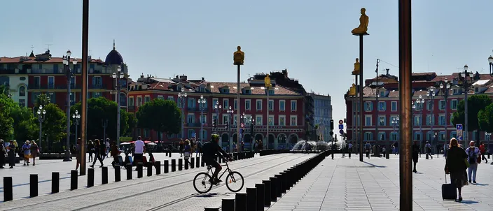 Place Masséna in Nice on a clear day: the wide pale paving of the square with a cyclist and pedestrians, tall poles carrying seated figures, and the red-ochre arcaded buildings along its far side.