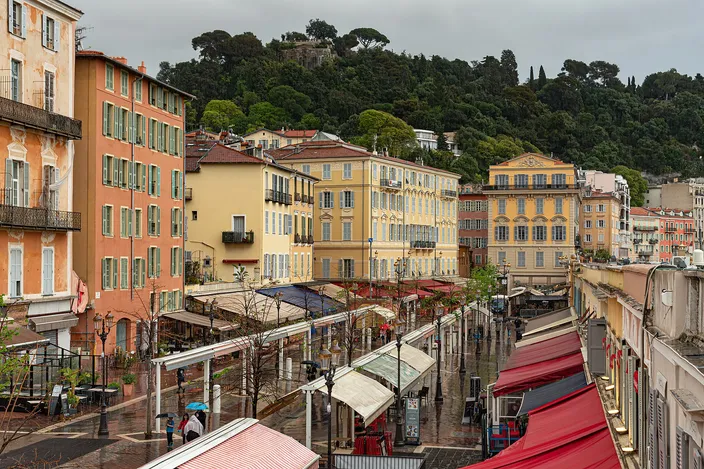 Cours Saleya in Nice's old town on a gray morning: the long market street lined with red and white awnings, ochre and yellow buildings, and the green slope of Castle Hill rising behind.